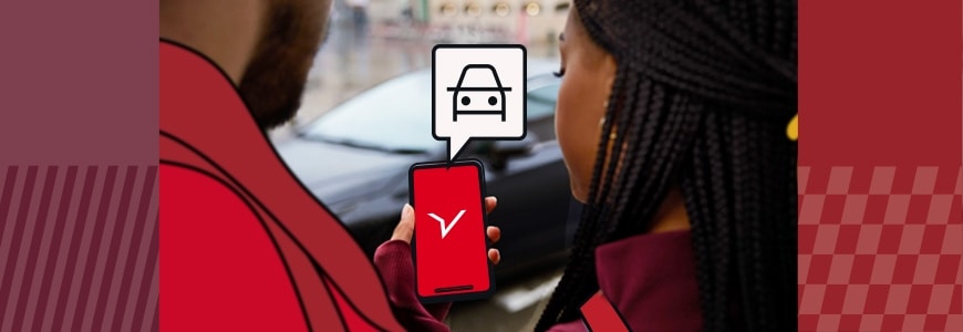 Due dipendenti in piedi davanti a un'auto del car sharing. Uno dei due ha in mano il proprio cellulare con il logo Freenow sullo schermo e l'icona di un'auto che spunta fuori.