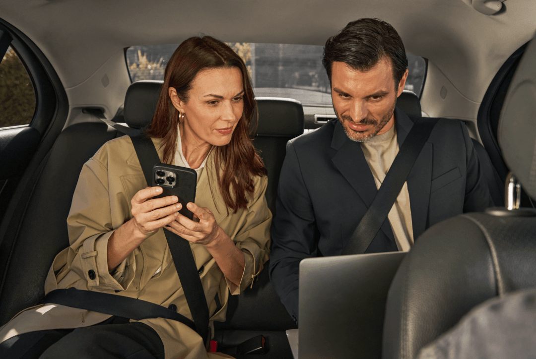 Due dipendenti in piedi con un taxi Freenow sullo sfondo. Un dipendente ha in mano il proprio cellulare con il logo di Freenow sullo schermo.